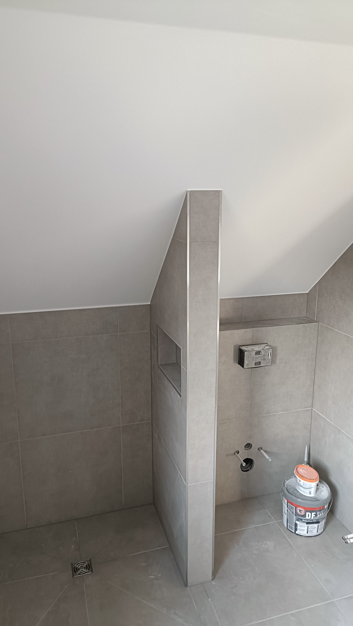 Wykończona łazienka na poddaszu z szarymi płytkami, widoczny odpływ liniowy i zabudowa WC. Na podłodze stoi wiadro z klejem do płytek.