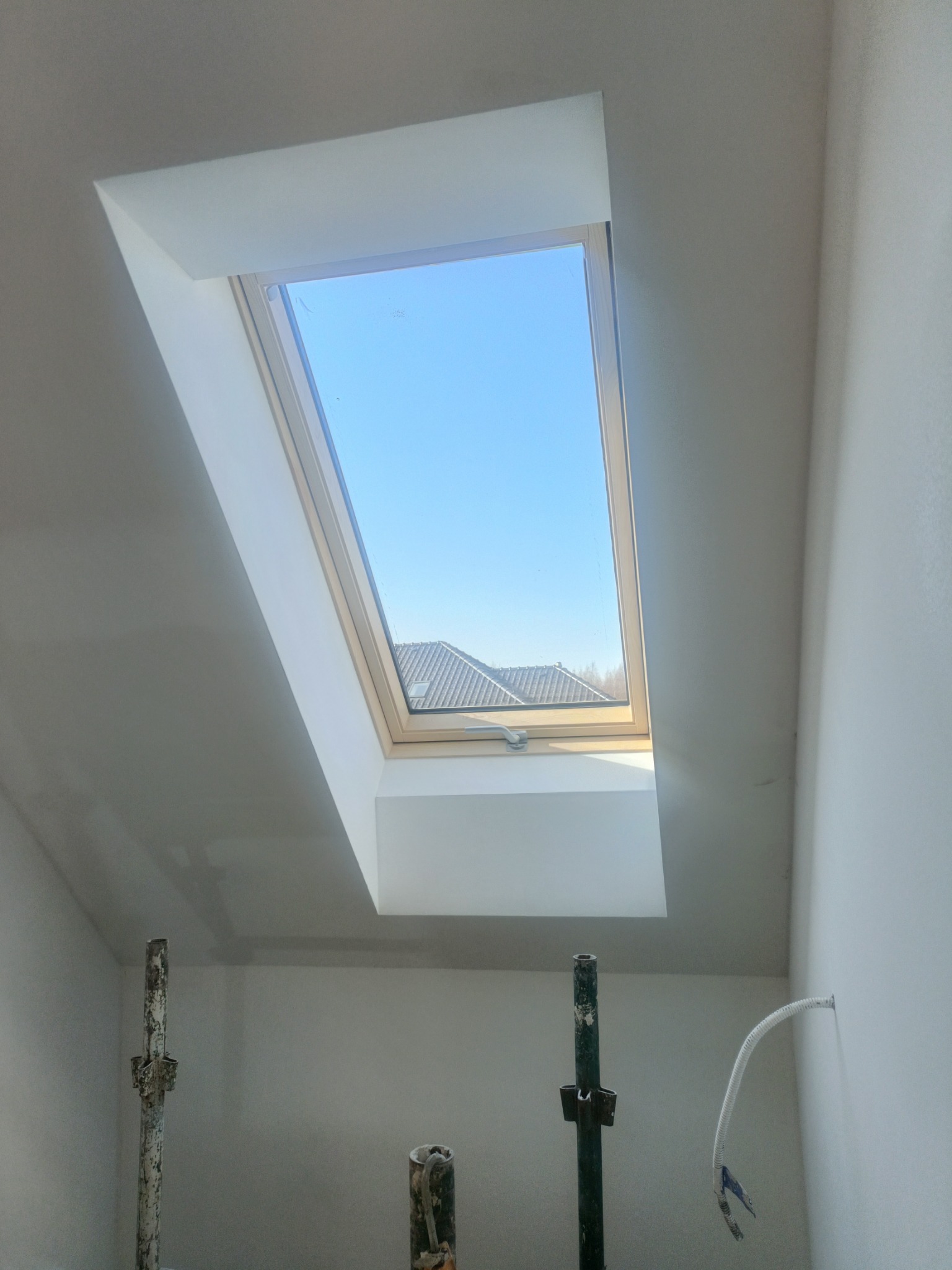 Wykończone obramowanie okna dachowego na poddaszu, widoczne rusztowania malarskie i wystający kabel. Świeżo pomalowane ściany i sufit w jasnym odcieniu.