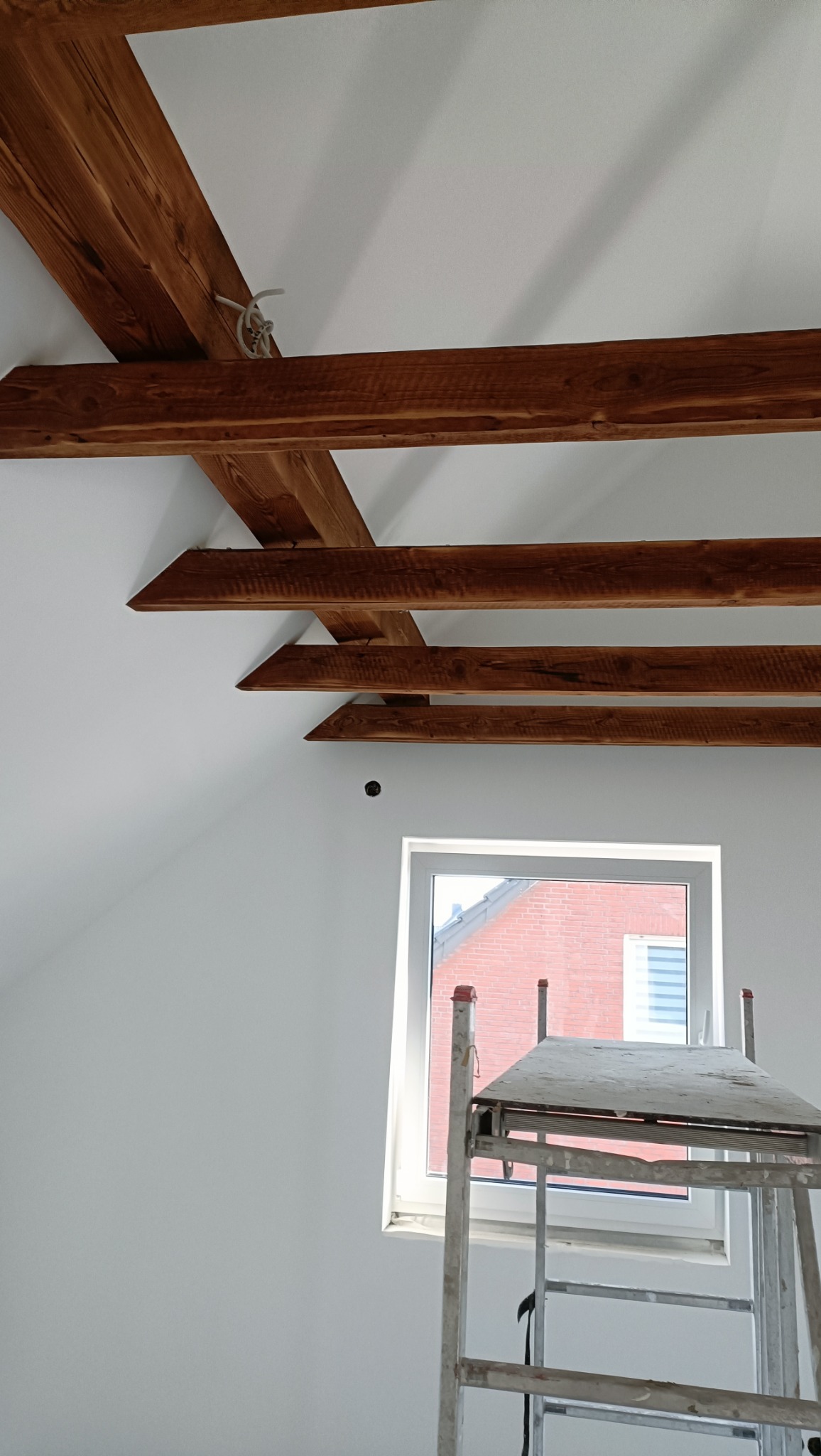 Białe poddasze z drewnianymi belkami stropowymi, widok na okno z rusztowaniem. Świeżo pomalowane ściany, surowe drewno belek. Wnętrze w trakcie prac wykończeniowych.