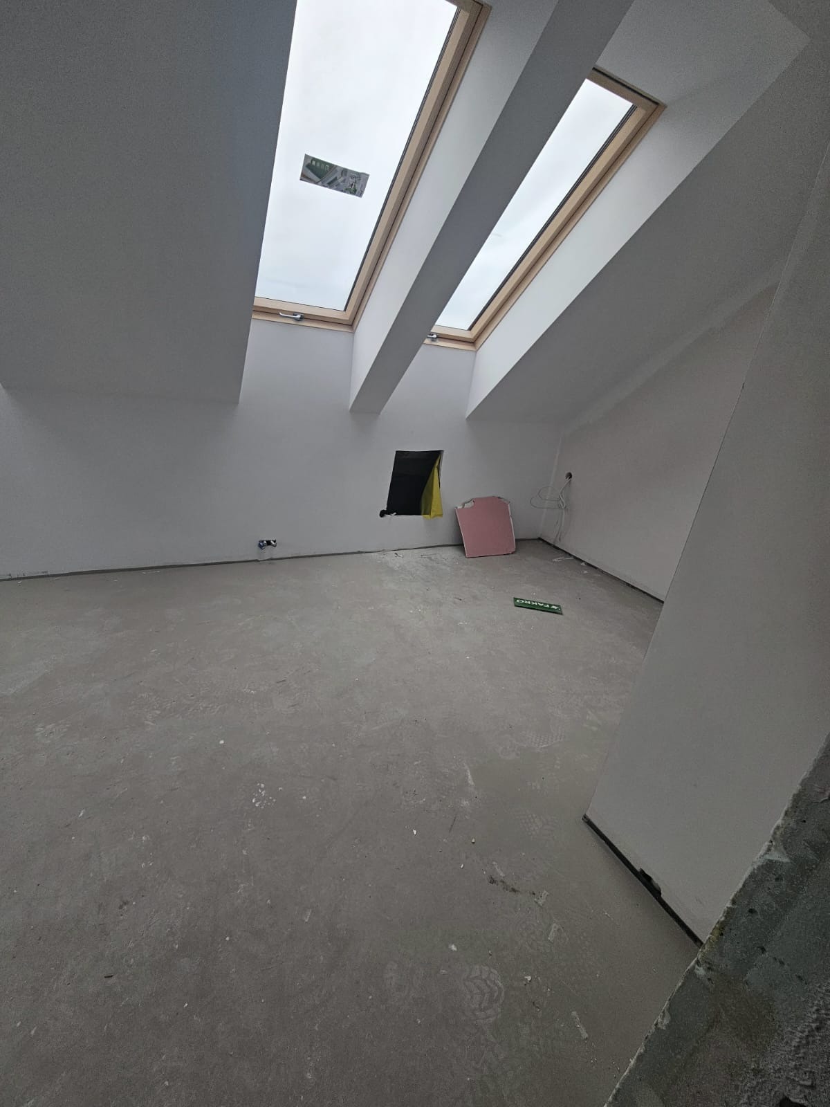 Poddasze po szpachlowaniu, z oknami dachowymi i przygotowanym otworem okiennym. Ściany pomalowane na biało, podłoga betonowa. Widoczne ślady po pracach.