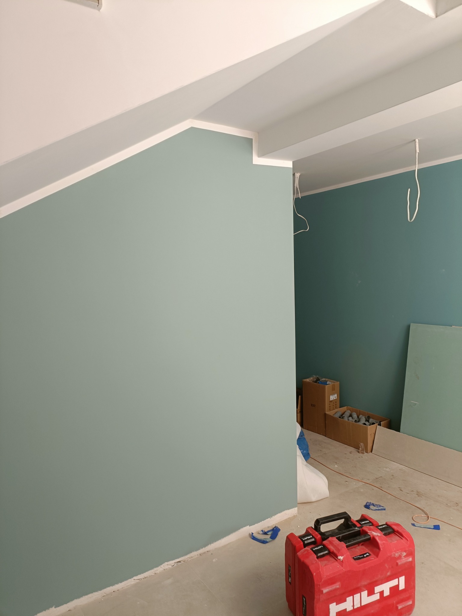Ściana pomalowana na dwa odcienie zieleni, z widoczną białą listwą sufitową i skrzynką narzędziową Hilti. Widoczne kable zwisające z sufitu.