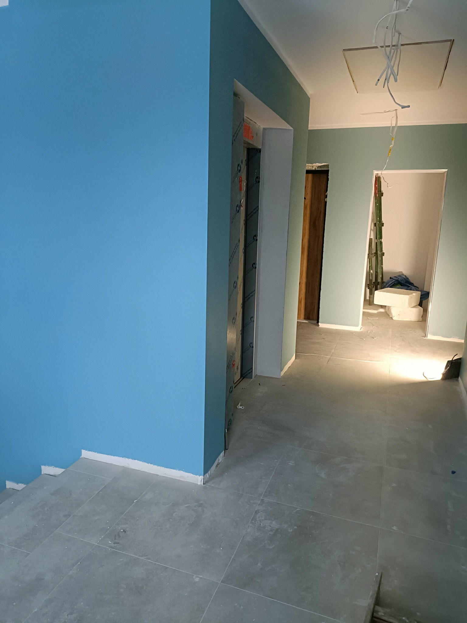 Świeżo pomalowane na błękitno ściany korytarza z szarą podłogą z płytek. Widoczne drzwi i instalacja elektryczna w suficie. Jasne, naturalne światło.