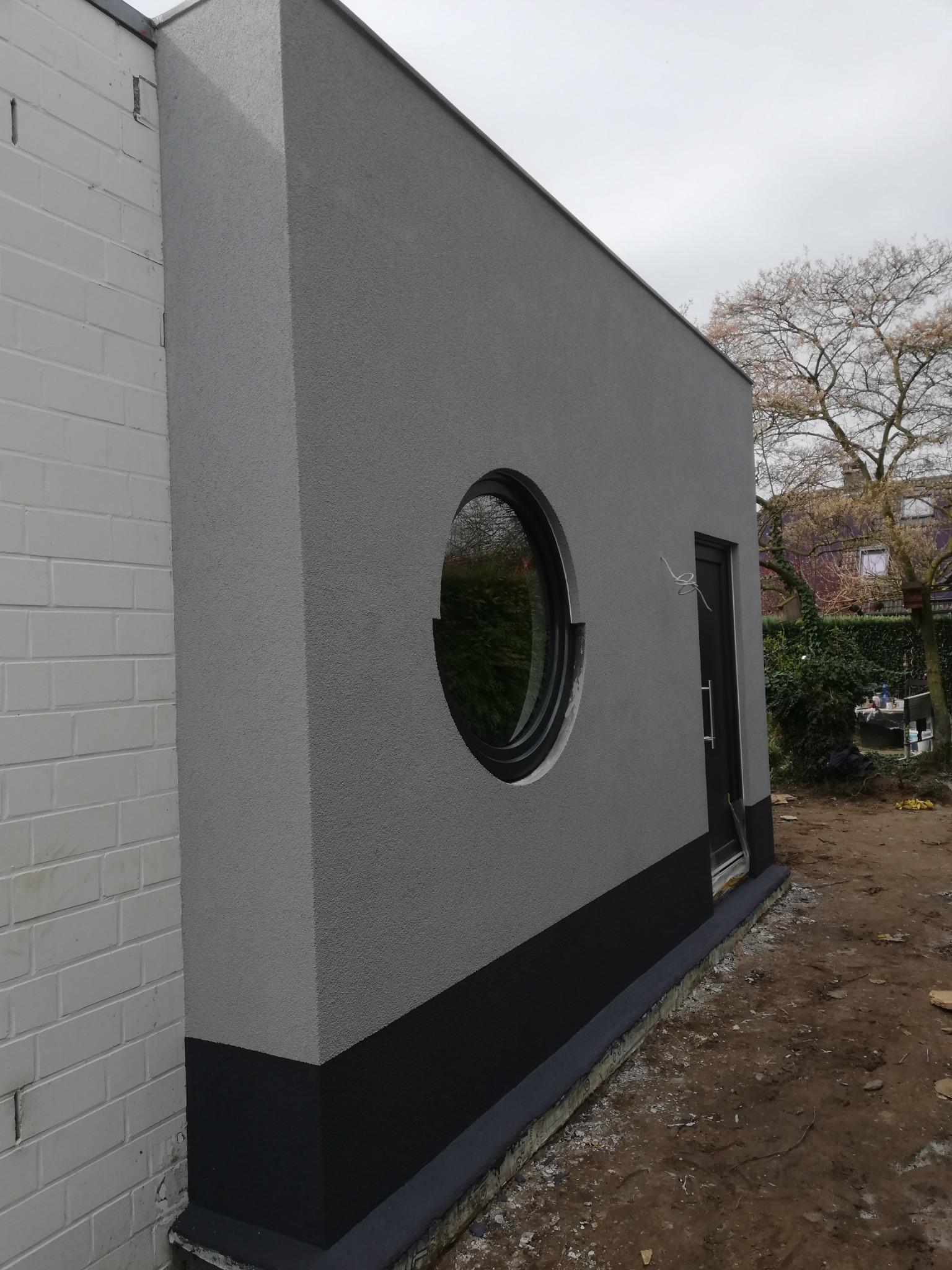 Szara elewacja budynku z okrągłym oknem i czarnym cokołem. Widoczny fragment białej ściany z cegły i drzewo w tle. Nowoczesny design.