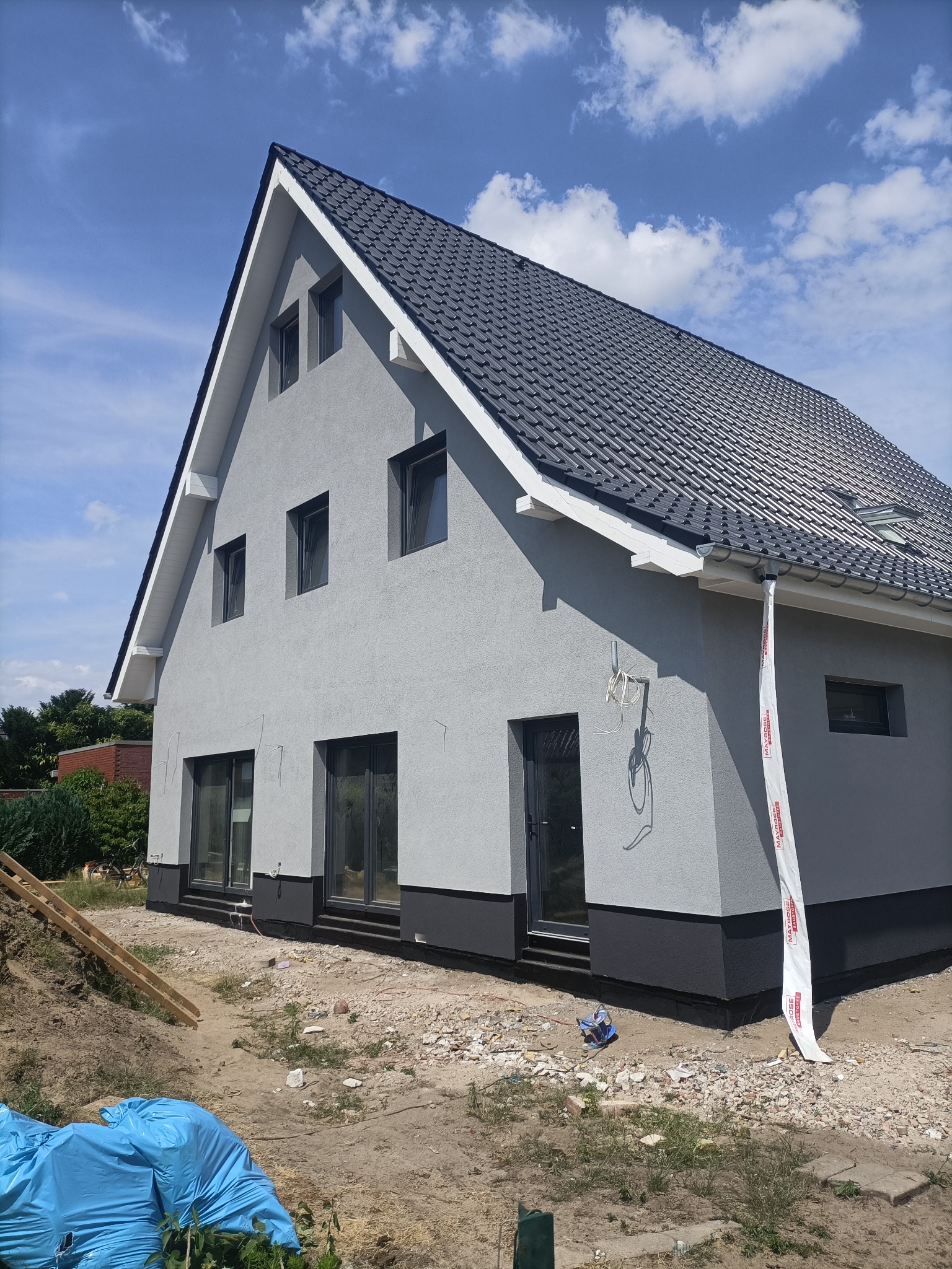 Dom jednorodzinny z szarą elewacją i czarną podmurówką, widok z boku, fragment rusztowania, błękitne niebo z chmurami w tle. Nowoczesny design.