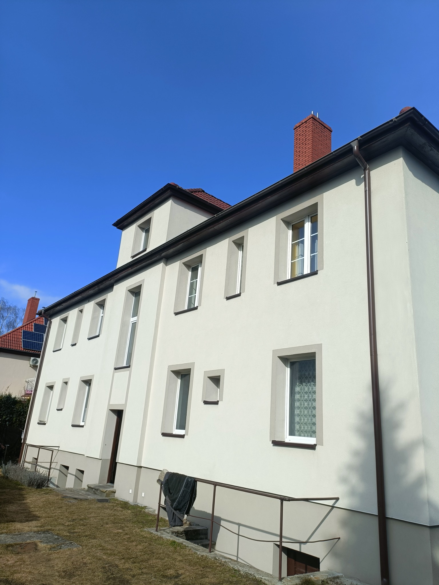 Odnowiona elewacja dwupiętrowego budynku z jasną fasadą, brązowymi rynnami i białymi oknami. Widoczne detale architektoniczne i zadbany teren wokół.