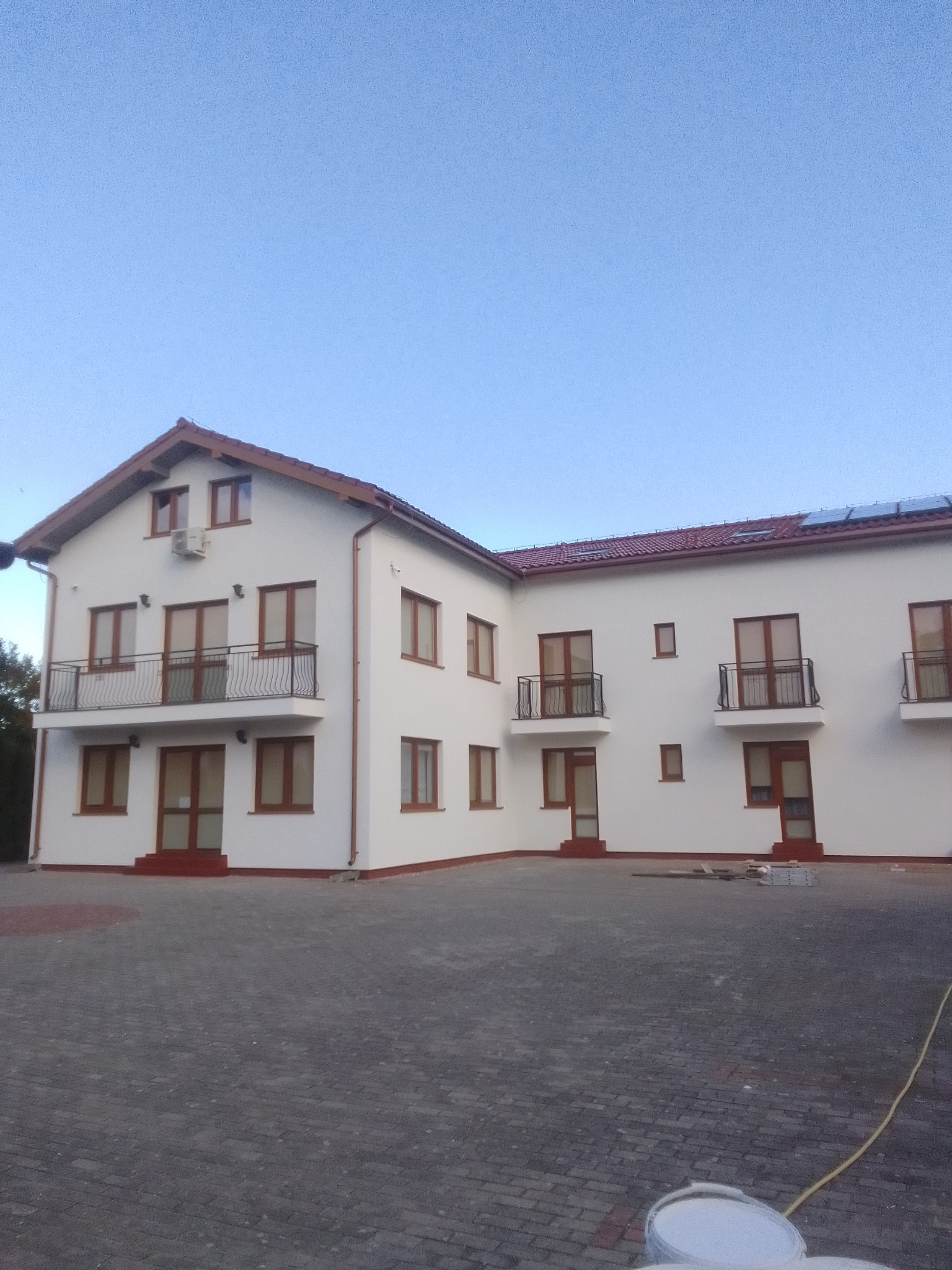 Biały budynek z brązowymi oknami i balkonami, widok z zewnątrz. Świeżo pomalowana elewacja, widoczne puszki z farbą na brukowanym podjeździe.