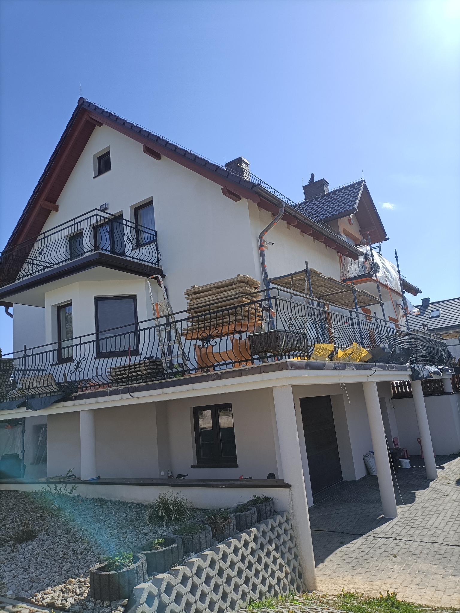 Dwupiętrowy dom z balkonami w trakcie prac elewacyjnych. Widoczne rusztowania, materiały budowlane i świeżo pomalowana fasada. Słoneczny dzień, błękitne niebo.