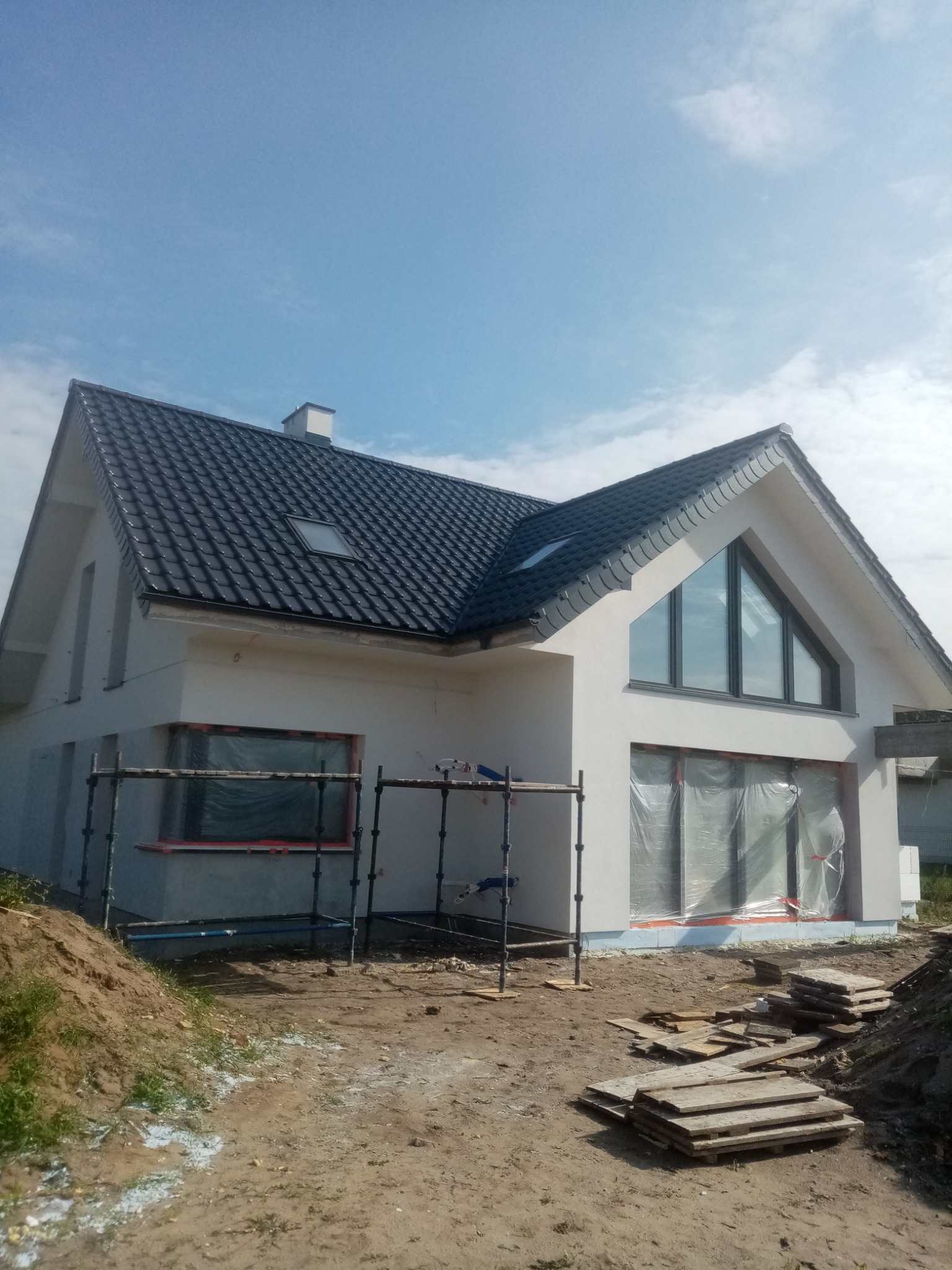 Nowoczesny dom jednorodzinny z ciemną dachówką i jasną elewacją w trakcie wykończenia. Widoczne rusztowania i zabezpieczone okna folią.