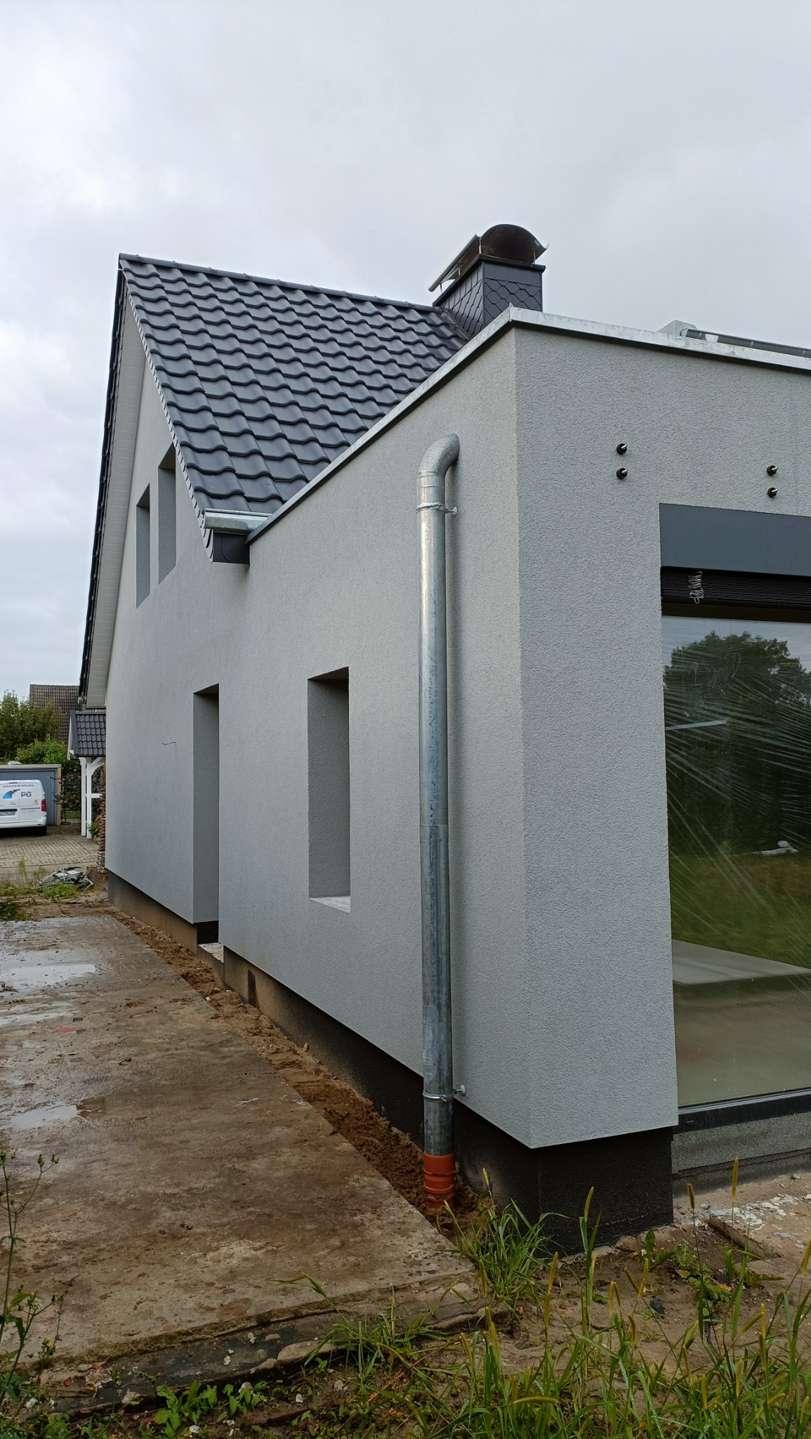 Szara elewacja domu jednorodzinnego z widoczną rynną spustową i fragmentem dachu z czarną dachówką. Nowoczesny styl, surowy krajobraz wokół budynku.