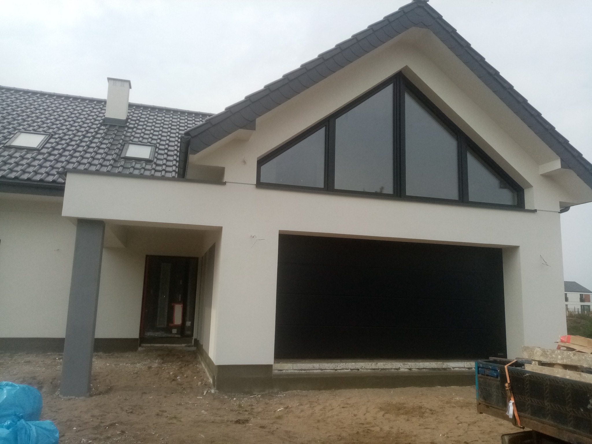 Nowoczesny dom jednorodzinny z garażem i dużym trójkątnym oknem na poddaszu. Biała elewacja, grafitowa dachówka i brama garażowa. Teren wokół domu w trakcie prac wykończeniowych.