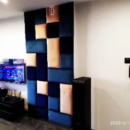 Panel ścienny z tapicerowanych kwadratów w odcieniach granatu, błękitu i beżu, stanowiący element dekoracyjny w nowoczesnym salonie z telewizorem i czarnymi meblami.