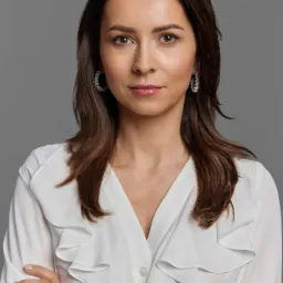 Portret kobiety z brązowymi włosami i białej bluzce z falbanami, skrzyżowane ręce, na szarym tle.