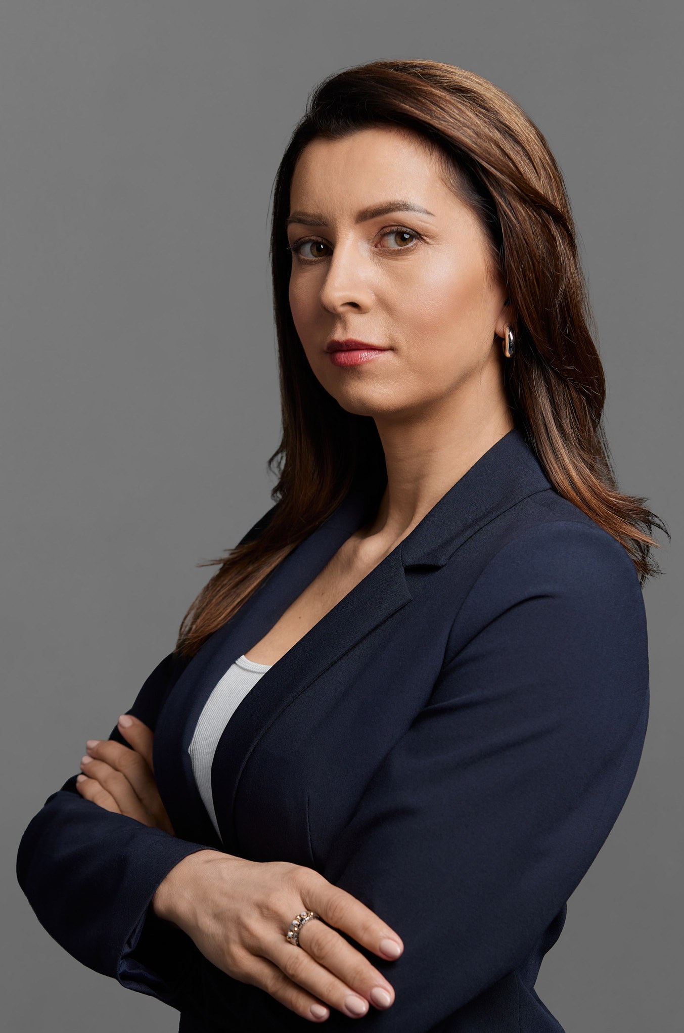 Portret kobiety w granatowej marynarce z rękami skrzyżowanymi, patrzącej wprost, z delikatnym uśmiechem i pierścionkiem na palcu, na jednolitym szarym tle.