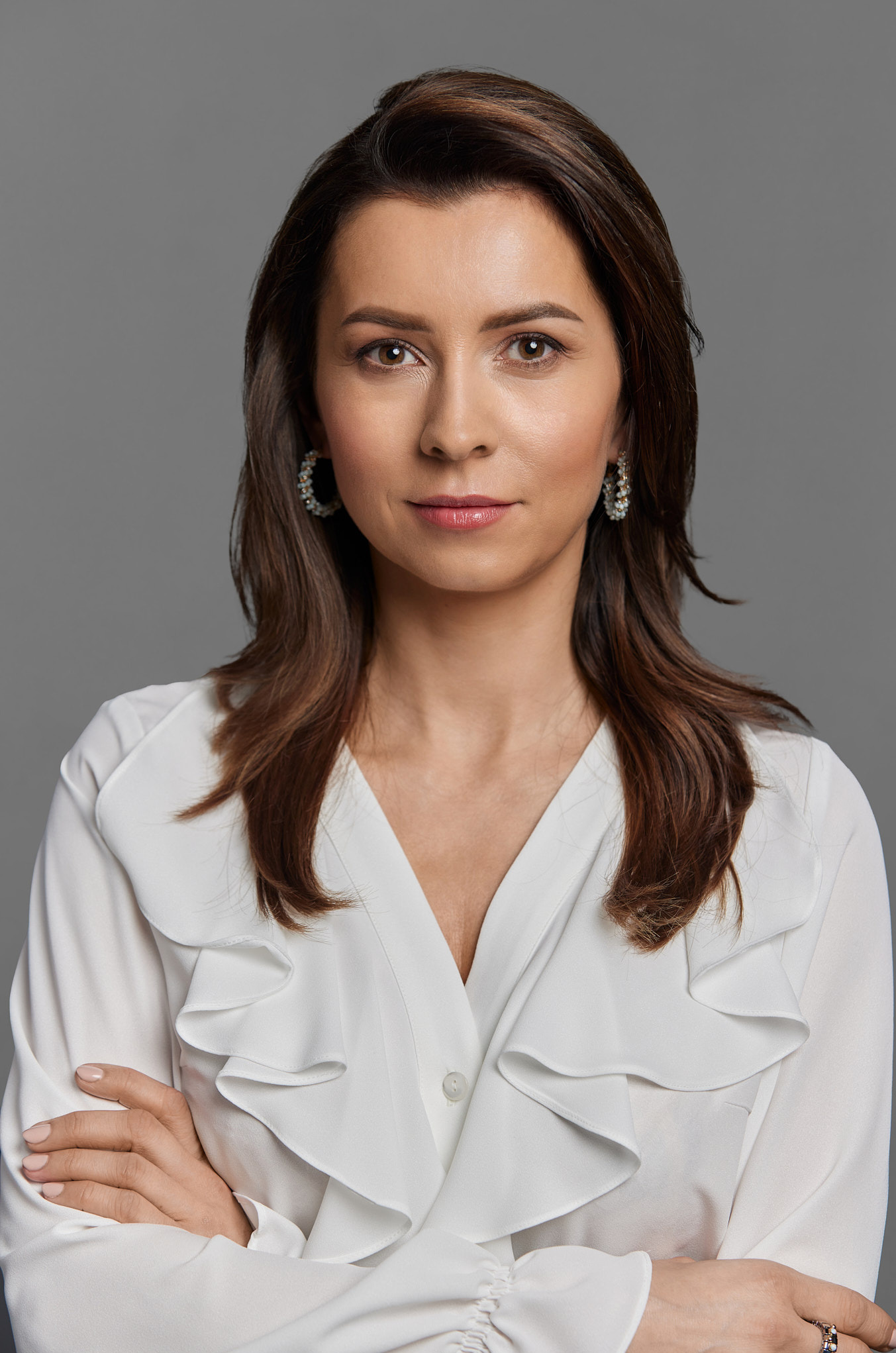 Portret kobiety z brązowymi włosami i białej bluzce z falbanami, skrzyżowane ręce, na szarym tle.