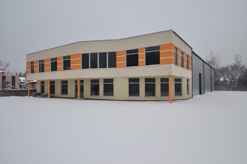 Dwukondygnacyjny budynek z częścią biurową i halą, pomalowany w odcieniach beżu z pomarańczowymi akcentami, otoczony śniegiem w pochmurny dzień.