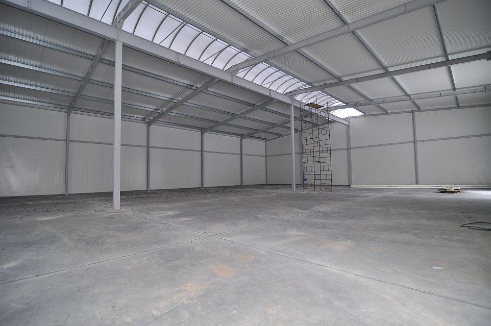 Wnętrze nowo wybudowanej hali z widoczną stalową konstrukcją, ścianami z płyt warstwowych i betonową posadzką, oświetloną przez świetliki dachowe.