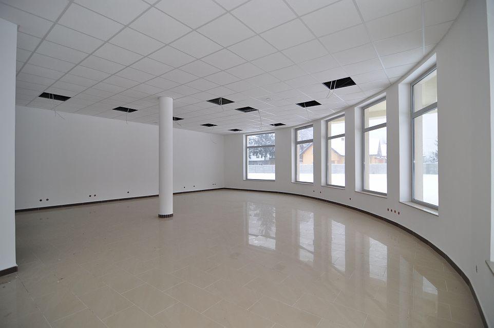 Wnętrze pustej hali z kolumną nośną, jasną podłogą z płytek i rzędem okien z widokiem na zabudowania mieszkalne.