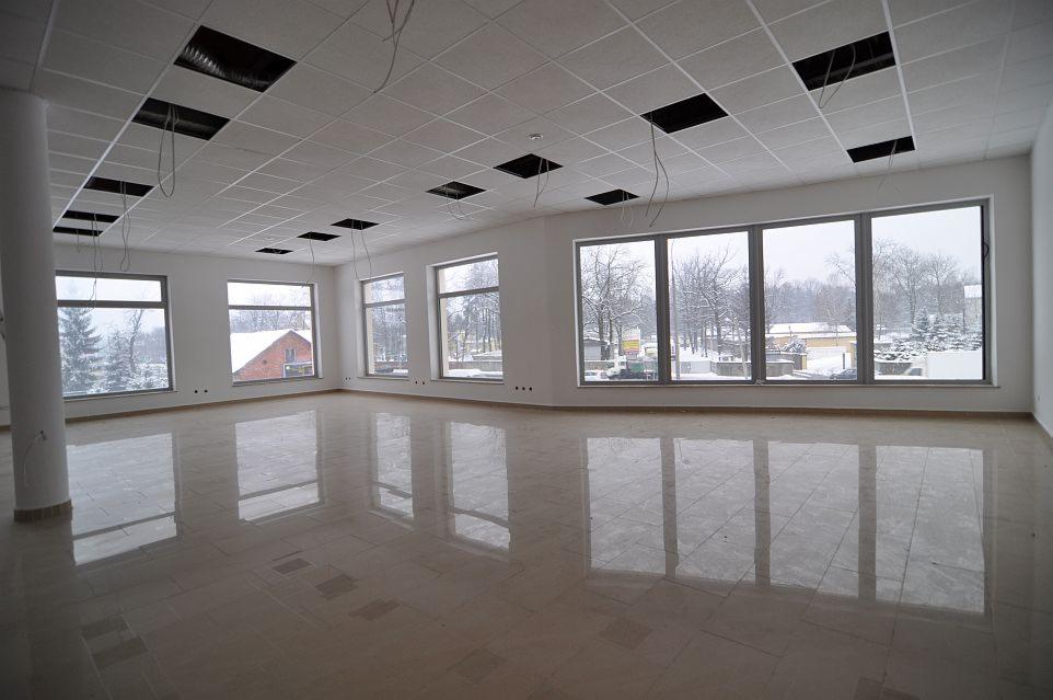 Wnętrze hali w stanie deweloperskim z widocznymi oknami, kolumną nośną i podwieszanym sufitem z otworami na instalacje.