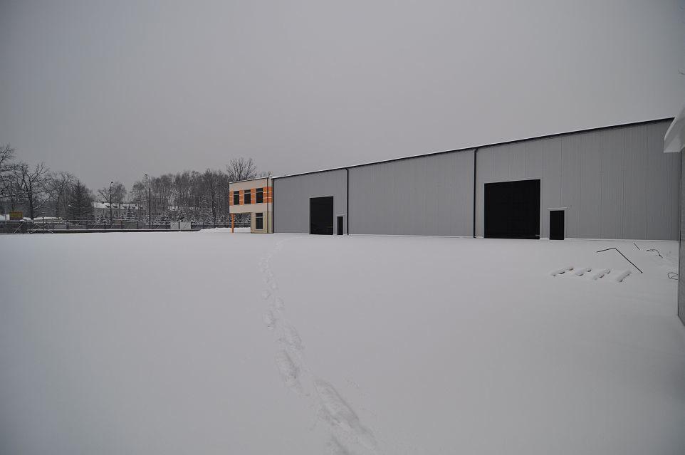 Zimowy krajobraz z widokiem na szarą halę z dwoma dużymi czarnymi bramami i dobudówką biurową z pomarańczowymi akcentami, pokrytą śniegiem, z widocznymi śladami stóp na śniegu prowadzącymi w stronę...