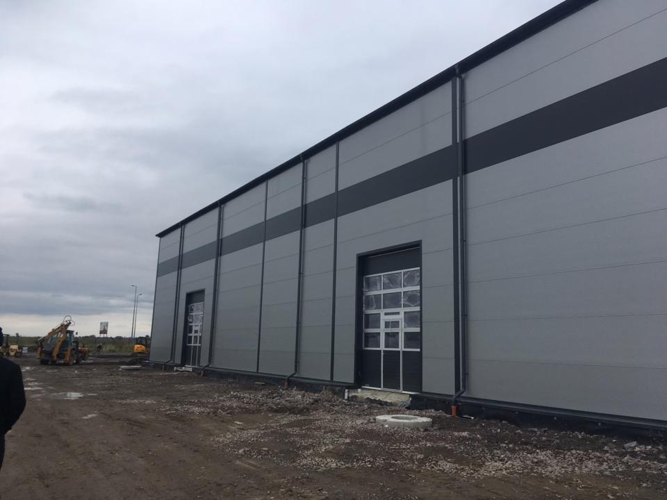 Szara hala przemysłowa z płyt warstwowych z dwoma bramami z folią ochronną na szybach, widoczny teren budowy i sprzęt budowlany w tle, pochmurne niebo.