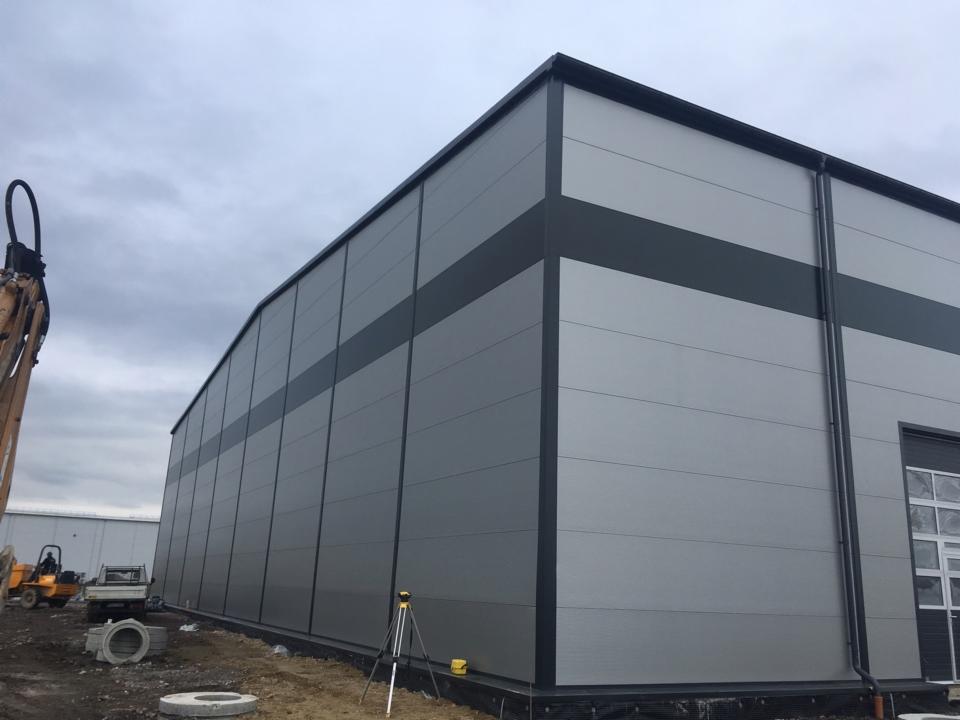 Szara hala przemysłowa z paneli warstwowych z ciemnym pasem, widoczny sprzęt budowlany i niwelator laserowy na zewnątrz.