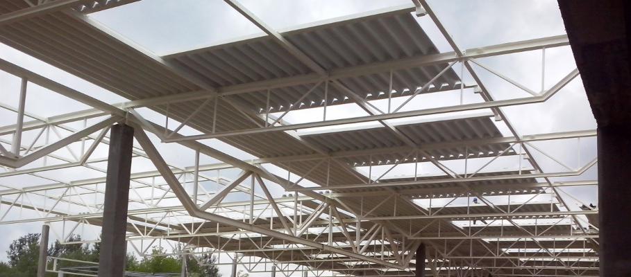 Wnętrze hali w budowie, widoczne stalowe elementy konstrukcyjne i częściowo zamontowane płyty warstwowe pod dachem, widok z dołu w pochmurny dzień.