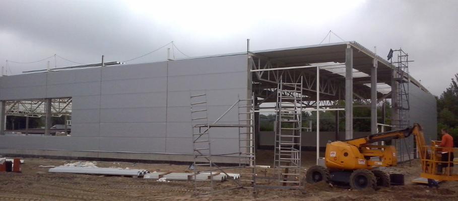 Wznoszona hala przemysłowa z szarej płyty warstwowej, widoczna stalowa konstrukcja szkieletowa, podnośnik koszowy i rusztowania na placu budowy w pochmurny dzień.