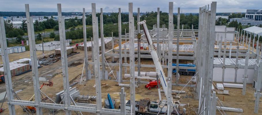 Wysoka konstrukcja hali w trakcie budowy, widoczne betonowe słupy i stalowe belki, na placu budowy pracuje dźwig, w tle drzewa i budynki.