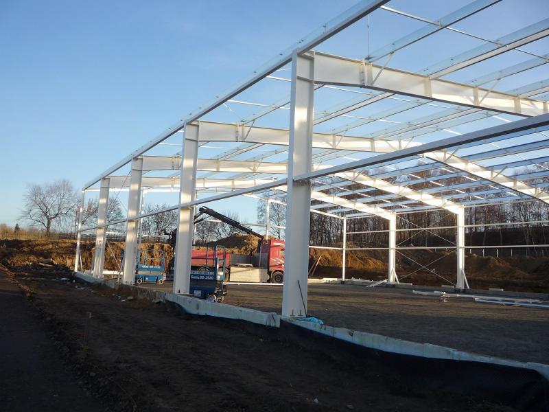 Wznoszenie szkieletu hali stalowej z widocznym dźwigiem i podnośnikami koszowymi na placu budowy w słoneczny dzień.