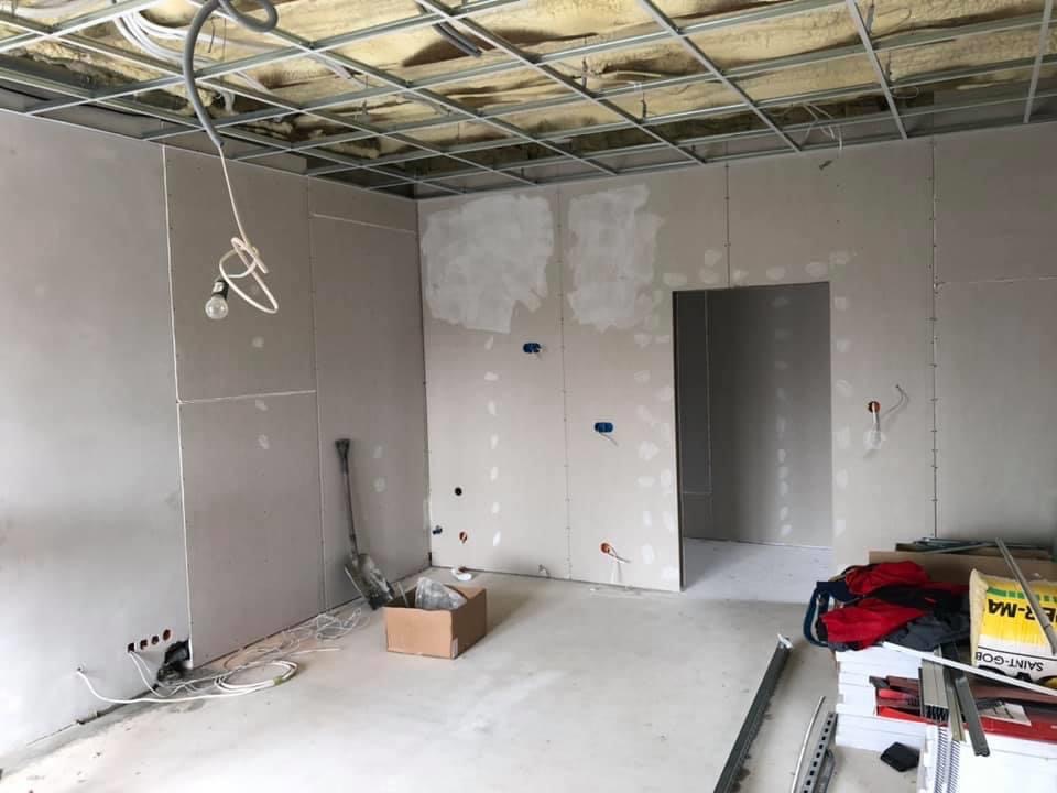 Wnętrze w trakcie montażu płyt kartonowo-gipsowych, widoczne profile sufitowe, częściowo wyszpachlowane ściany z widocznymi otworami pod instalacje elektryczne, karton i narzędzia na podłodze.