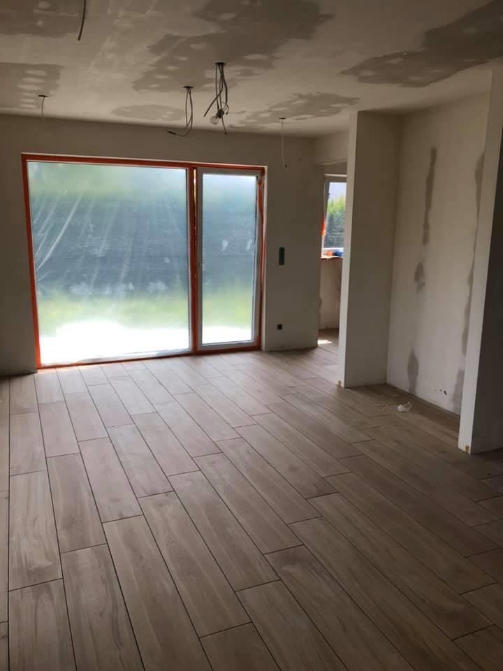 Wnętrze w trakcie remontu: położona podłoga z płytek imitujących drewno, okno zabezpieczone folią, ściany częściowo wyszpachlowane, widoczne kable zwisające z sufitu.