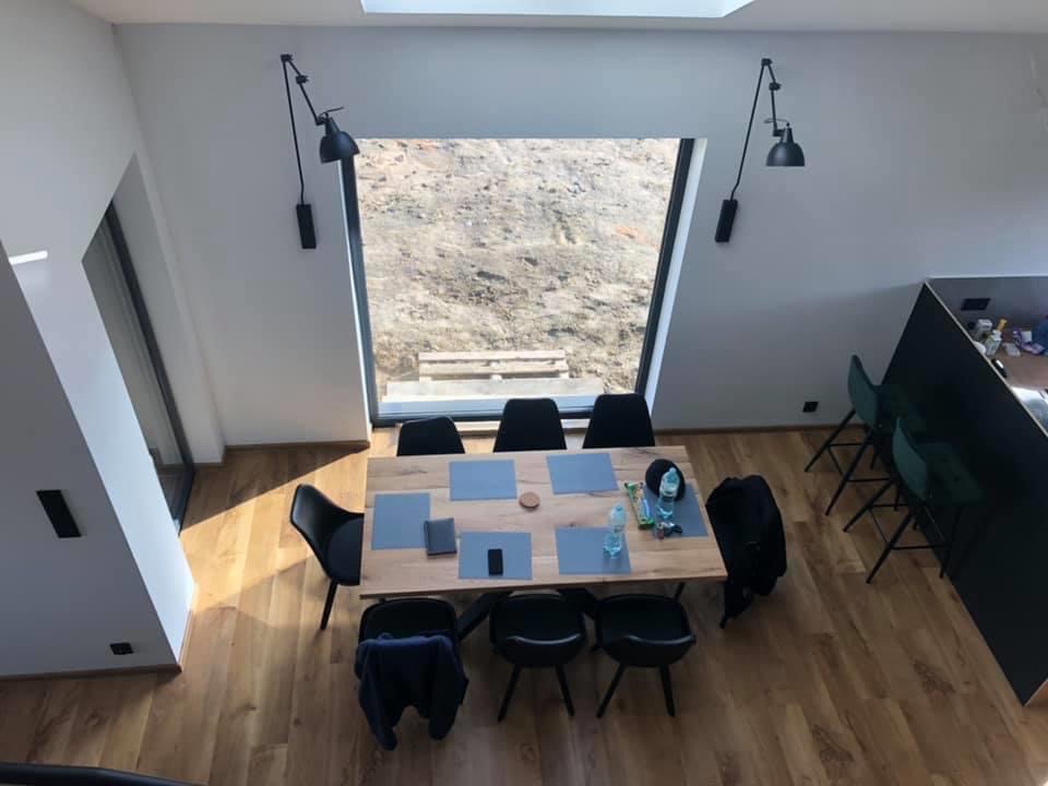 Wnętrze nowoczesnego domu z drewnianą podłogą i stołem jadalnym, widok z góry na minimalistyczną aranżację z czarnymi lampami ściennymi i dużym oknem z widokiem na ziemię.