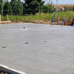 Zakład Ogólnobudowlany Michał Tomalak TOM-MAX - Wylana, gładka płyta fundamentowa na tle zieleni i błękitnego nieba. Widoczne wystające rury instalacyjne. Świeży beton, brązowa ziemia wokół.