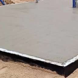 Zakład Ogólnobudowlany Michał Tomalak TOM-MAX - Świeżo wylany fundament z widocznymi instalacjami, otoczony piaskiem i drzewami. Ujęcie z bliska, ukazujące gładką powierzchnię betonu i detale przygotowania pod budowę.