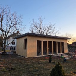 Zakład Ogólnobudowlany Michał Tomalak TOM-MAX - Konstrukcja szkieletowa budynku gospodarczego z płyt OSB, widoczna na tle domu jednorodzinnego i drzew bez liści, w promieniach zachodzącego słońca.