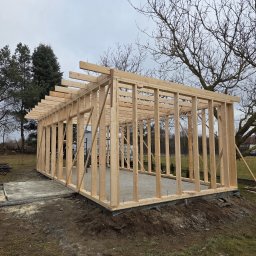 Zakład Ogólnobudowlany Michał Tomalak TOM-MAX - Drewniany szkielet konstrukcji budynku w trakcie budowy, widoczne słupy i belki, fundament częściowo wylany, drzewa w tle, pochmurne niebo.