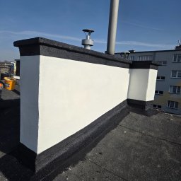 Zakład Ogólnobudowlany Michał Tomalak TOM-MAX - Odnawianie komina na dachu pokrytym papą: świeżo pomalowana, biała ściana komina z czarnym wykończeniem, widok z góry na tle błękitnego nieba i budynku.