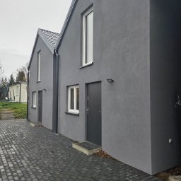 Zakład Ogólnobudowlany Michał Tomalak TOM-MAX - Dwa identyczne, szare domy z ciemnymi dachami i kostką brukową. Minimalistyczny styl, nowoczesne oświetlenie zewnętrzne, białe ramy okienne. Pochmurne niebo w tle.