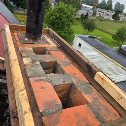 Zakład Ogólnobudowlany Michał Tomalak TOM-MAX - Wznoszenie komina z cegły na dachu, widoczne drewniane wzmocnienia i czarny przewód kominowy. W tle budynki i zieleń, pochmurne niebo.