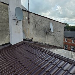 Zakład Ogólnobudowlany Michał Tomalak TOM-MAX - Nowy, brązowy dach z blachy trapezowej, widok z góry. Dwie anteny satelitarne na tle budynku z szarą elewacją i fragmentem nieba z chmurami.