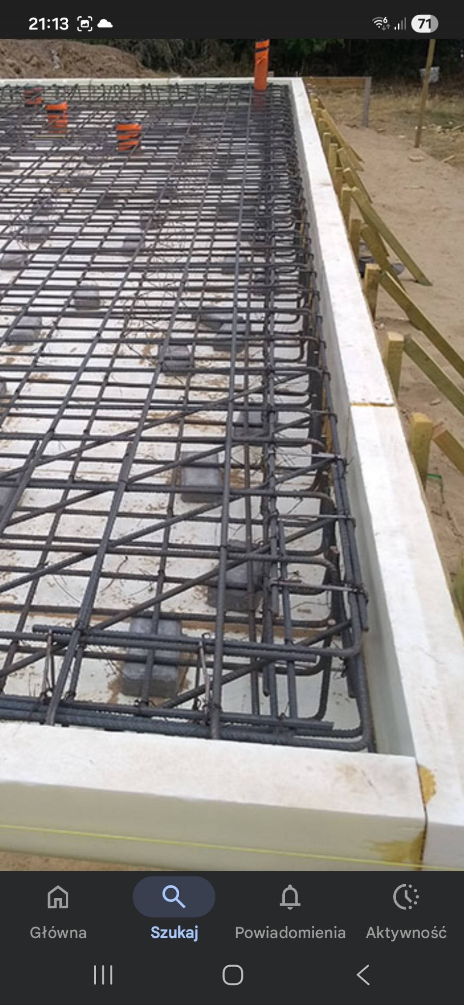 Zbrojenie fundamentów w szalunku z białego styropianu. Widoczne pręty zbrojeniowe ułożone w kratę i pomarańczowe rury instalacyjne. Drewniane podpory szalunku.