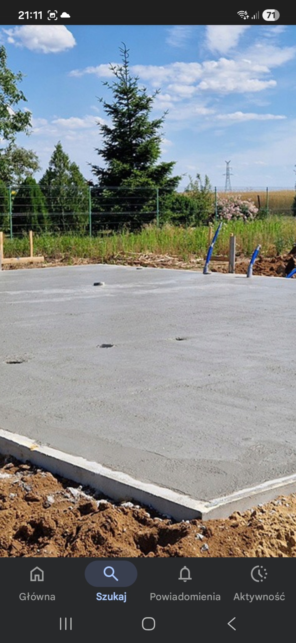 Wylana, gładka płyta fundamentowa na tle zieleni i błękitnego nieba. Widoczne wystające rury instalacyjne. Świeży beton, brązowa ziemia wokół.