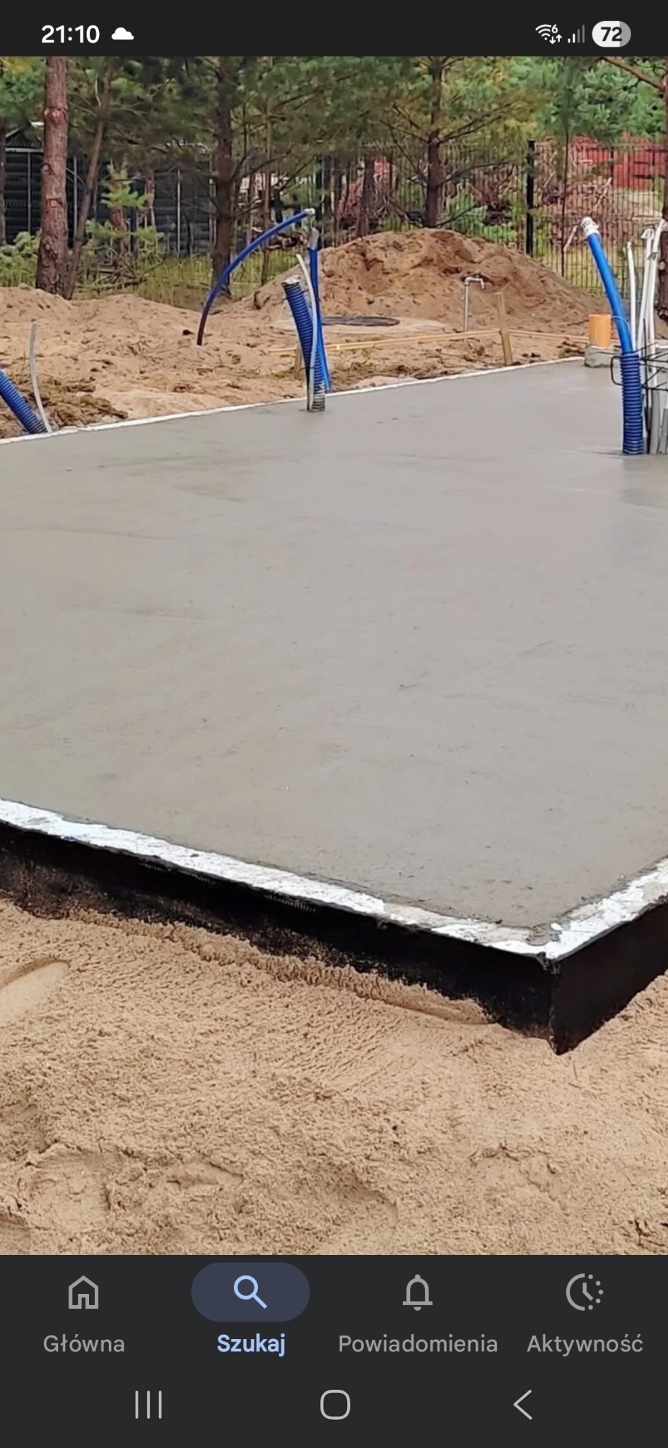 Świeżo wylany fundament z widocznymi instalacjami, otoczony piaskiem i drzewami. Ujęcie z bliska, ukazujące gładką powierzchnię betonu i detale przygotowania pod budowę.