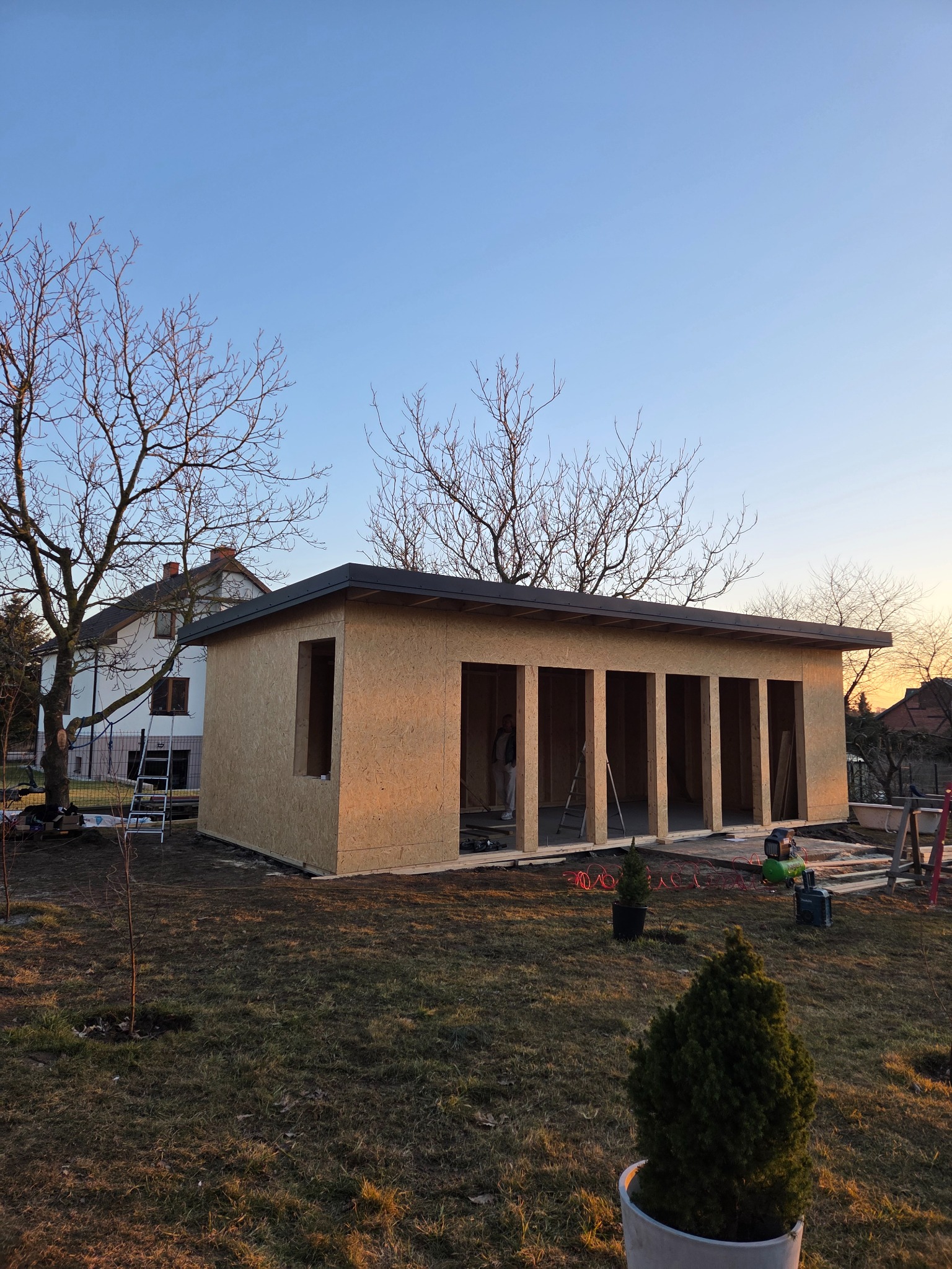 Konstrukcja szkieletowa budynku gospodarczego z płyt OSB, widoczna na tle domu jednorodzinnego i drzew bez liści, w promieniach zachodzącego słońca.