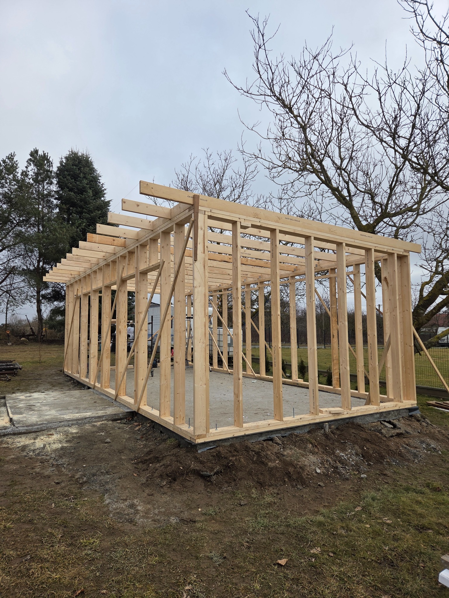 Drewniany szkielet konstrukcji budynku w trakcie budowy, widoczne słupy i belki, fundament częściowo wylany, drzewa w tle, pochmurne niebo.