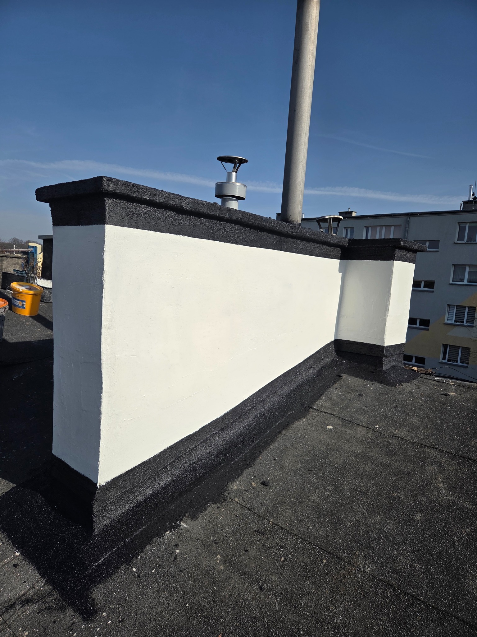 Odnawianie komina na dachu pokrytym papą: świeżo pomalowana, biała ściana komina z czarnym wykończeniem, widok z góry na tle błękitnego nieba i budynku.