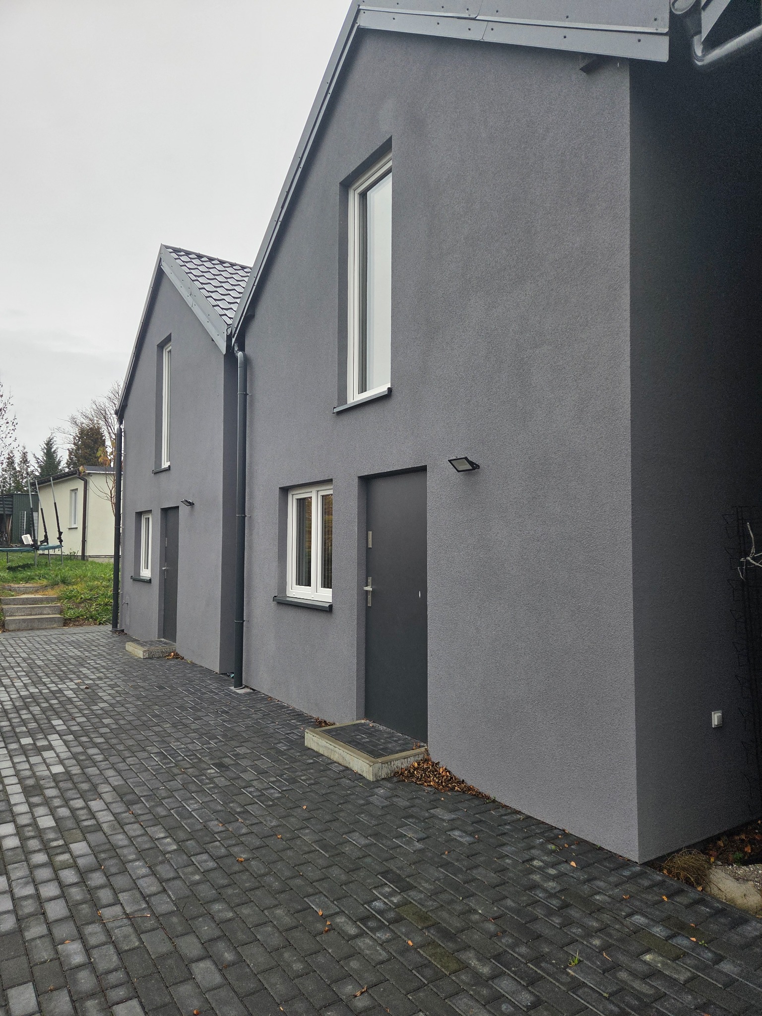 Dwa identyczne, szare domy z ciemnymi dachami i kostką brukową. Minimalistyczny styl, nowoczesne oświetlenie zewnętrzne, białe ramy okienne. Pochmurne niebo w tle.