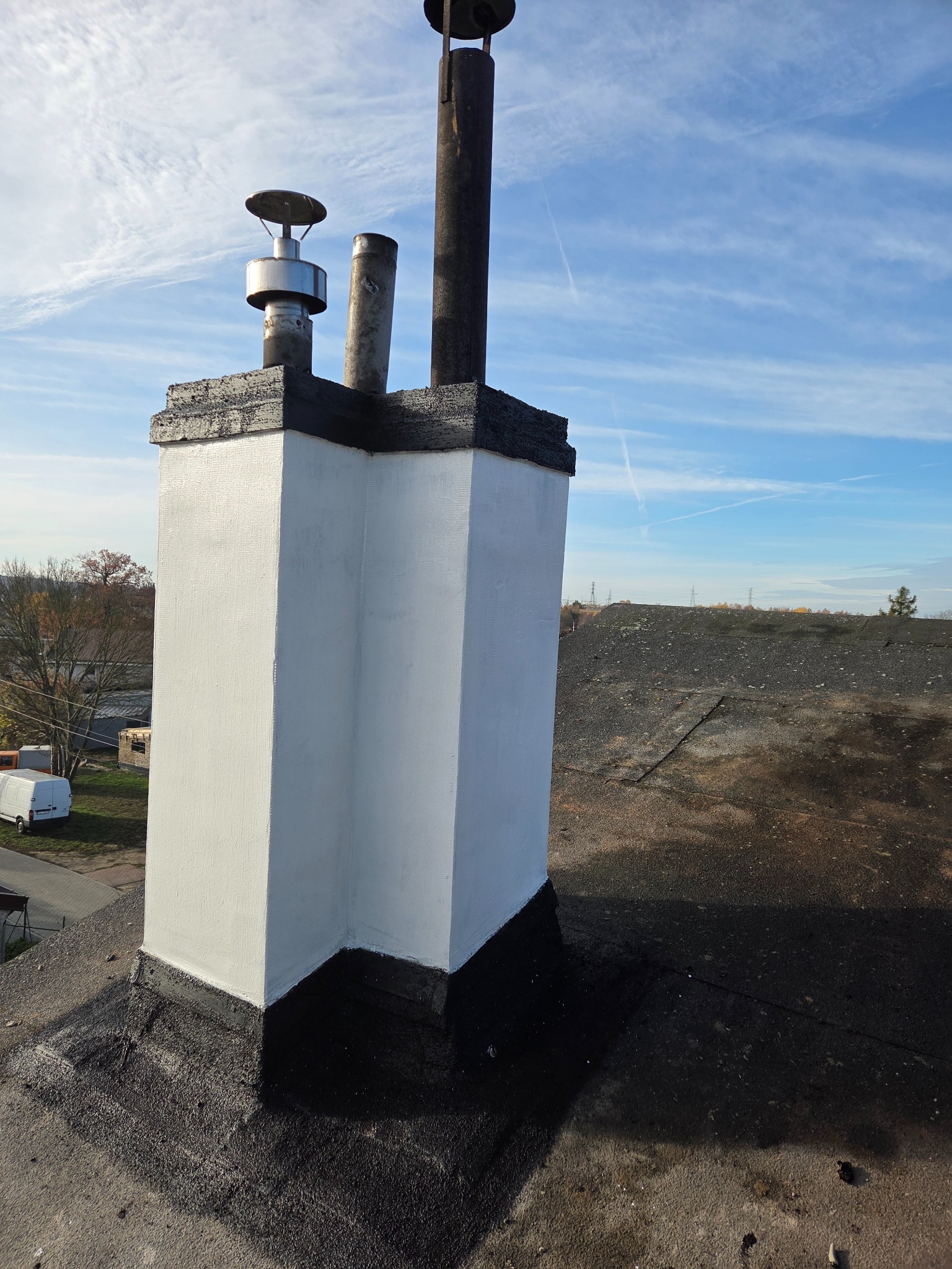 Biały, odnowiony komin na dachu pokrytym papą, z widocznymi elementami wentylacyjnymi i fragmentem nieba z chmurami. Komin pomalowany na biało, z czarnym wykończeniem.