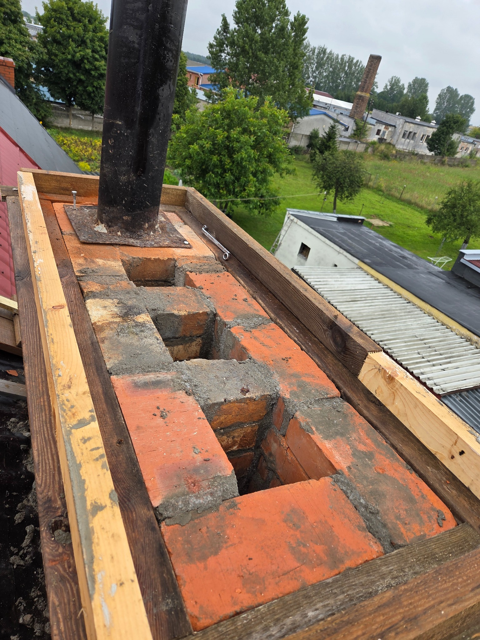 Wznoszenie komina z cegły na dachu, widoczne drewniane wzmocnienia i czarny przewód kominowy. W tle budynki i zieleń, pochmurne niebo.