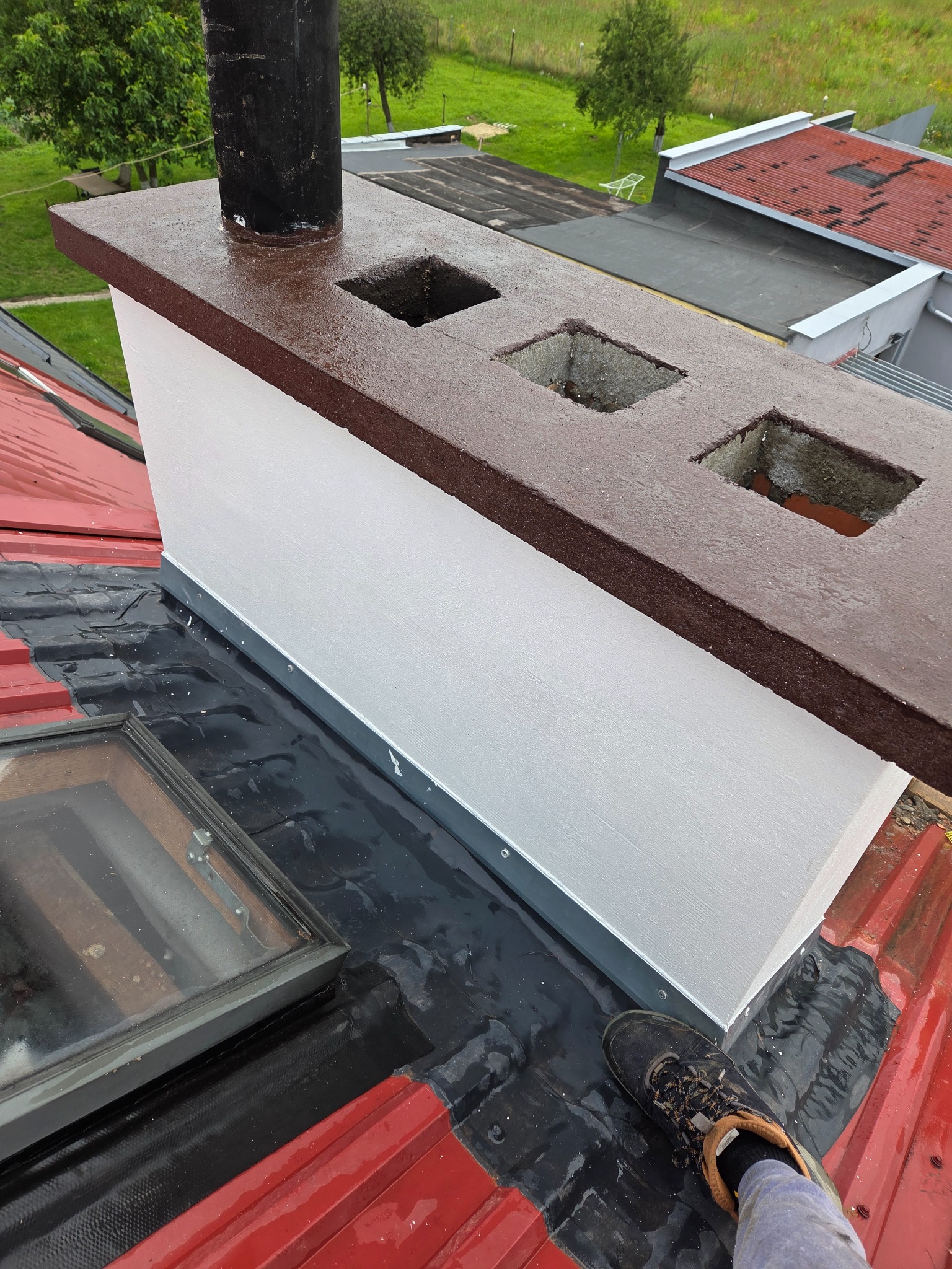 Odnawianie komina na dachu z czerwonej blachy trapezowej. Widoczne trzy otwory w brązowej obudowie komina i czarny przewód kominowy. Prace dekarskie w toku.